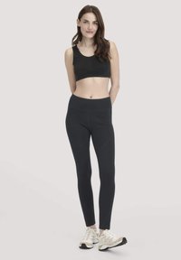 Vrouw die staat in een zwarte sportbeha, zwarte leggings en witte sneakers tegen een effen lichte achtergrond, glimlachend met haar handen achter haar rug.