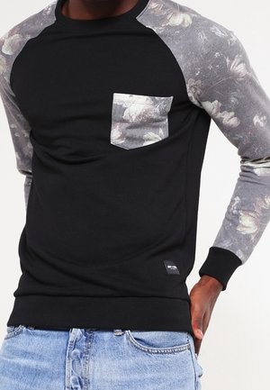 Czarna koszula z długim rękawem z szarymi rękawami w kwiatowy wzór oraz dopasowaną kieszenią na piersi, noszona z jasnoniebieskimi dżinsami.