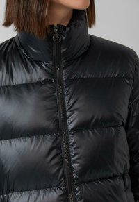 Melna pūkaina jaka ar augstu apkakli, rāvējslēdzēju un izšūtu dizainu. Audumam ir spīdīga tekstūra ar redzamām vīlēm.