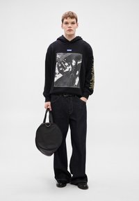 Μαύρο φούτερ με κουκούλα και εκτυπωμένο σχέδιο, συνδυασμένο με σκουρόχρωμο τζιν παντελόνι φαρδιάς γραμμής. Κρατάει μια μαύρη στρογγυλή τσάντα με λαβή στο πάνω μέρος.
