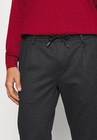 Pantalons gris à motifs avec taille élastique à cordon. Comprend un tissu texturé et des poches latérales. Associés à un pull rouge.