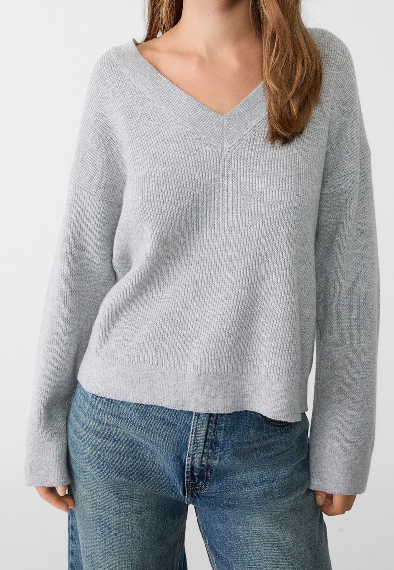 Femme portant un pull à col en V côtelé gris clair et un jean bleu, debout devant un fond blanc uni, montrant le haut du corps et les bras.