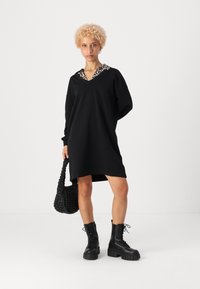 Črna pulover obleka z ovratnikom v leoprdjem vzorcu. Dolgi rokavi, sproščen kroj, v kombinaciji s črno tkanino torbo in masivnimi črnimi vojničkimi čevlji.