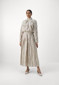 Rochie midi pliată din țesătură bej deschis, cu modele florale texturate, guler înalt și talie elastică; asortată cu pantofi cu toc în nuanțe deschise.