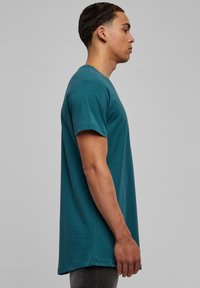 Tricou din bumbac de culoare teal, cu mâneci scurte, guler rotund și tăietură dreaptă, expus pe un model din lateral, pe un fundal gri.