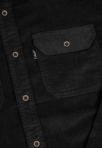 Schwarzes Cordshirt mit Knopfleiste, einem einzelnen Brusttasche, kontrastierenden kupferfarbenen Knöpfen und strukturiertem Stoff.