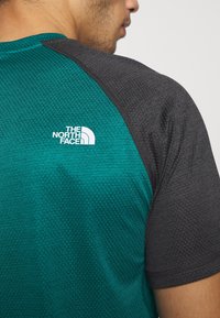 Koszulka sportowa z krótkim rękawem w kolorze zielonkawym z teksturowanej tkaniny i ciemnoszarymi rękawami. Biały logo znajduje się na klatce piersiowej.