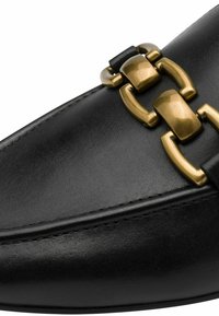 Svart läderloafer med en slät yta, som har en framträdande guldtonad kedjeaccent över framfoten och detaljerad sömnad längs kanterna.
