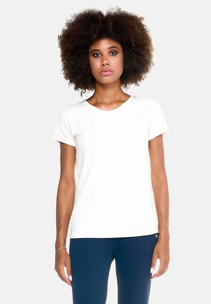 Femme aux cheveux bouclés portant un t-shirt blanc uni à manches courtes et un pantalon bleu foncé, debout devant un fond clair uni.