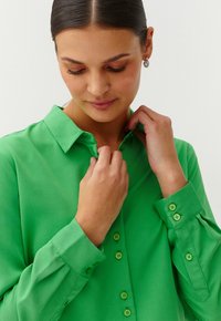 Donna con capelli scuri in camicia verde con bottoni che sistema il colletto, indossando orecchini a cerchio in argento, su uno sfondo chiaro e semplice.
