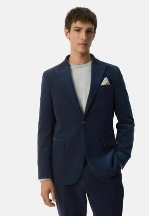 Ung mand iført en navyblå fløjlsdragt med en hvid lommetørklæde og en lysegrå trøje med rund hals, poserer mod en ensfarvet baggrund.