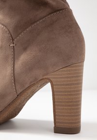 Stivaletto alto in suede marrone con tacco in legno texture. Presenta dettagli cuciti e una superficie liscia e morbida.