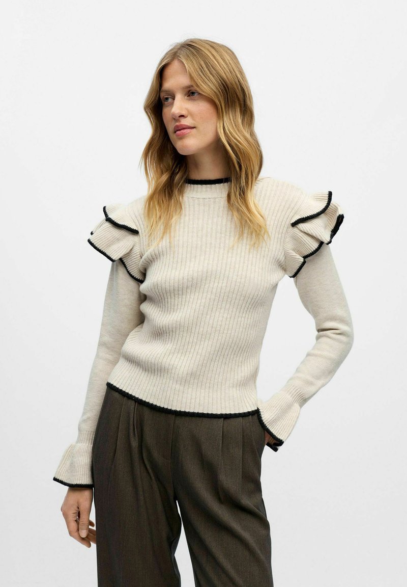 Pull à côtes beige avec des bordures noires, manches évasées à volants et coupe ajustée, associé à un pantalon plissé gris.