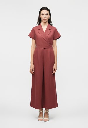 Femeie purtând o salopetă cu mâneci scurte, de culoare cărămizie, cu pantaloni evazați și guler în stil înfășurat, asortată cu sandale albe cu barete.