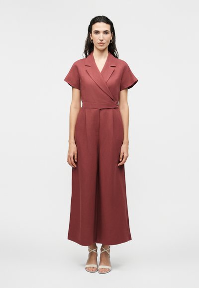 Femme portant une combinaison de couleur rouille à manches courtes, pantalon évasé et col cache-cœur, associée à des sandales blanches à brides.