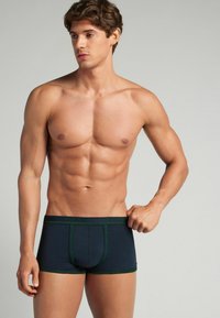 Mörkblå boxerkalsonger med grön söm, tillverkade av ett mjukt, elastiskt material. Har en åtsittande passform och en bekväm midjeresår.