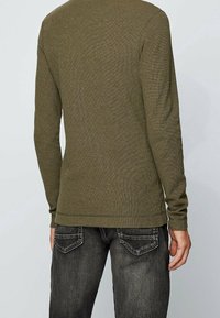 Oliwkowa koszula z długim rękawem o strukturalnym splocie, dopasowanym kroju i okrągłym dekolcie, w zestawie z ciemnoszarymi dżinsami.