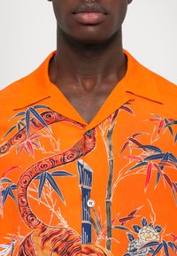 Lühikese varrukaga oranž särk, millel on detailne lille- ja bambusikujundus sinise, punase ja smaragdrohelise värviga. Sile kangas ja nööbilukuga.