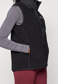Black fleece vest with a high collar, featuring a zippered front, side pockets, and gray long-sleeve top underneath. Soft, textured material.