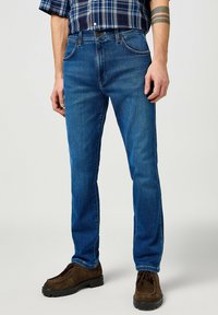 Jeans de denim azul en un corte recto clásico, con cinco bolsillos, costuras ligeras y un acabado ligeramente desvaído. Combinados con zapatos de ante marrones.