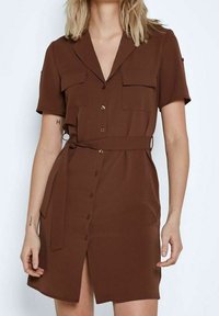 Hnědé šaty s krátkým rukávem vyrobené z hladké látky, s knoflíkovou légou vpředu, dvěma náprsními kapsami a zavázaným páskem v pase.