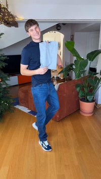 Maglietta in tessuto grigio tenuta in mano, abbinata a jeans blu. Elementi circostanti includono piante in vaso e un divano accogliente in una stanza ben illuminata.