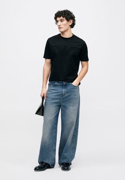 Μαύρο t-shirt με στρογγυλή λαιμόκοψη και ανάγλυφο λογότυπο, σε συνδυασμό με ανοιχτό μπλε τζιν παντελόνι φαρδιάς γραμμής και μαύρα παπούτσια. Κρατάει ένα μαύρο αξεσουάρ.