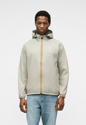 Mees, kes kannab helehallist kapuutsiga jopet triibulise luku ja helesiniste teksadega, seisab ühetaolise halli tausta ees.