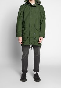 Grön parkajacka med huva, med framfickor, knapplås och dragsko i nederkant. Kombinerad med mörkgrå byxor och svarta sneakers.