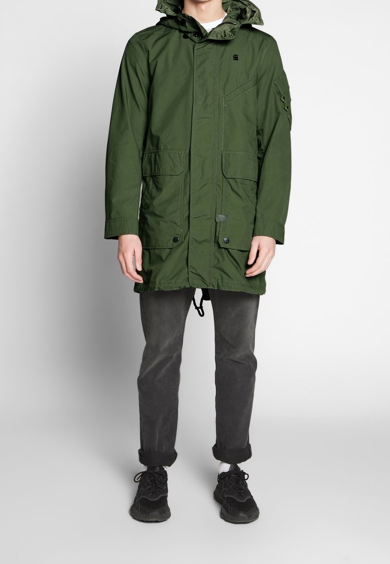 Grön parkajacka med huva, med framfickor, knapplås och dragsko i nederkant. Kombinerad med mörkgrå byxor och svarta sneakers.