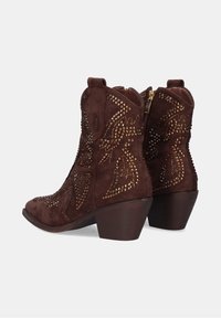 Botines de ante marrón con puntera afilada, adornados con tachuelas doradas en un patrón decorativo y un tacón bajo apilado. Cremallera en la parte trasera.
