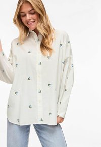 Jeune femme souriante, portant une chemise ample blanche à boutons avec de petites imprimés floraux bleus et un jean bleu clair, sur un fond uni.