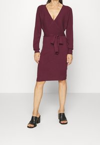 Burgundy omlottklänning med långa ärmar, v-ringad hals och midjeband. Tillverkad av ribbat stickat tyg, med knä-lång längd. Svarta klacktofflor.