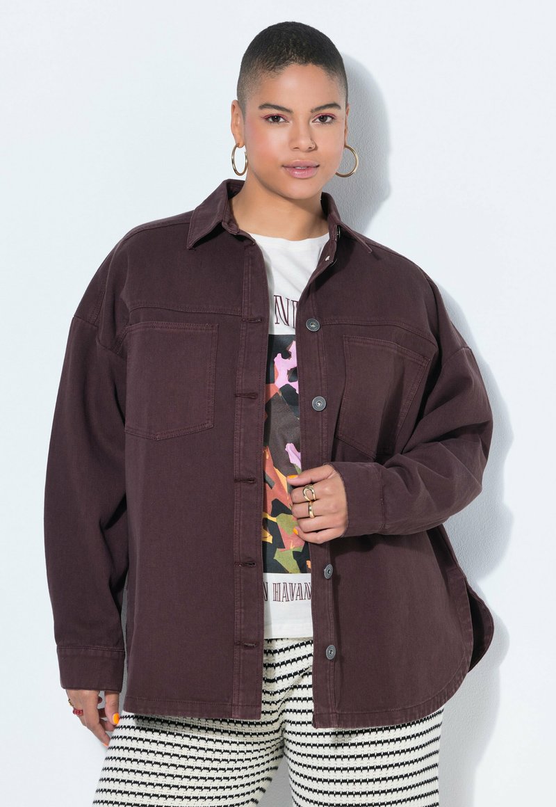 Person mit kurzen Haaren trägt Creolen, eine übergroße dunkelbraune Jacke, ein weißes T-Shirt mit Grafik und gestreifte Hose, posiert vor weißem Hintergrund.