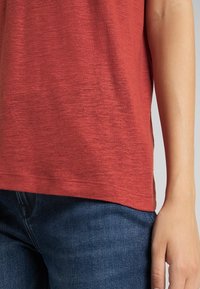 Gros plan sur une personne portant une chemise rouge à manches courtes et un jean bleu, avec le bras droit reposant le long du corps.