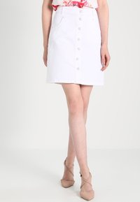 Jupe en denim blanc en A avec fermeture à boutons sur le devant, deux poches latérales et coutures renforcées. Le tissu a une texture lisse.