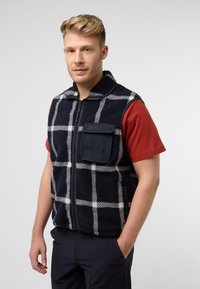 Fleece-Weste mit navy- und cremefarbenem Karomuster, durchgehendem Reißverschluss und einer Brusttasche. Das Modell trägt ein rotes T-Shirt darunter und dunkle Hosen.