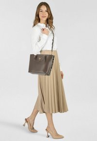 Bolso marrón estructurado con acabado suave, doble asa y correa estampada. Modelo lleva una camisa blanca abotonada y una falda beige plisada.