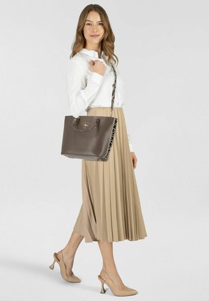Structured brown handbag with a smooth finish, dual handles, and a patterned strap. Model wears a white button-up and pleated beige skirt.