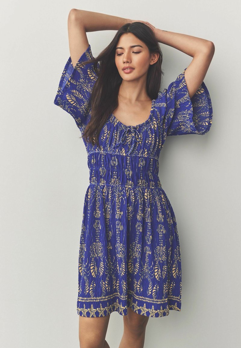 Robe bleue avec des manches courtes et larges ; présente un motif de feuilles dorées et une taille froncée. Le tissu semble léger et texturé.