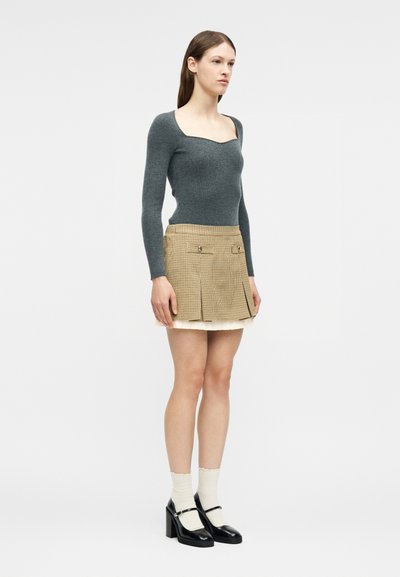 Femme debout portant un haut gris à manches longues, une mini-jupe plissée beige, des chaussettes blanches et des talons Mary Jane noirs sur un fond clair uni.