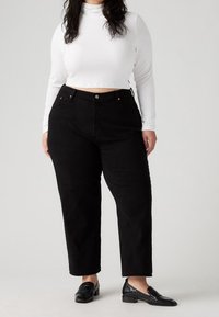 Pantalones de mezclilla negros de tiro alto con corte recto, que presentan un cierre de botón y sutiles acentos en cobre. Combinados con un cuello alto de canalé blanco.