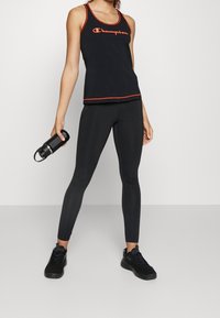 Svart topp utan ärmar med röd kant och "Champion"-logotyp, kombinerad med svarta leggings. Modellen håller en svart vattenflaska och bär svarta sneakers.