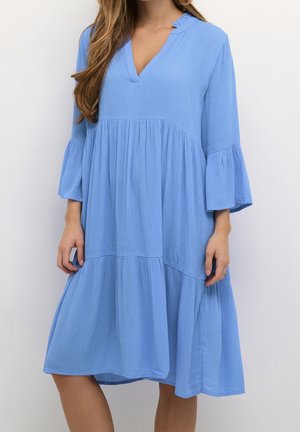 Frau trägt ein locker sitzendes, knielanges hellblaues Kleid mit dreiviertel Glockenärmeln und V-Ausschnitt.