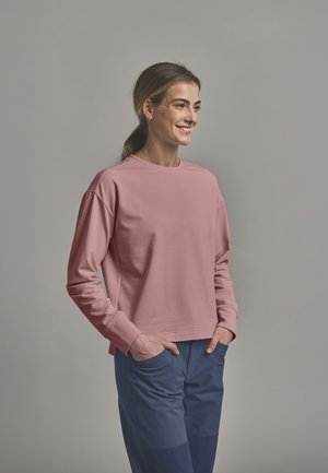 Frau lächelt mit Händen in den Taschen, trägt ein langärmliges staubrosa Oberteil und marineblaue Hosen, stehend vor einem einfachen grauen Hintergrund.