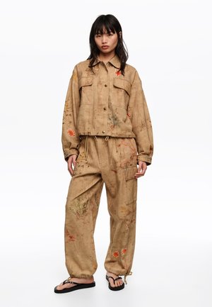 Kobieta ubrana w luźną, beżową kurtkę i spodnie w kwiatowe wzory, stojąca boso na białym tle.