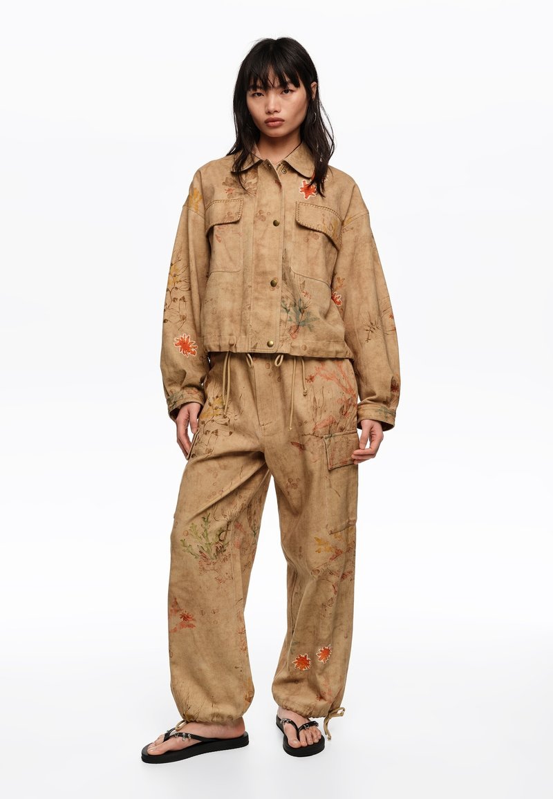 Woman wearing loose tan jacket and pants with floral prints, standing barefoot on a white background.