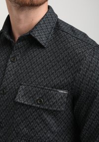 Donker, gestreept shirt met een chevronpatroon in zwart- en grijstinten. Bevat knopen, een kraag en een borstzak met een merklabel.