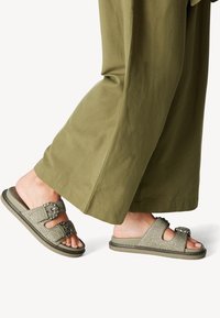Platta sandaler med två breda tygremmar, olivgrön färg, texturerad vävdetalj och dekorativa spännen; sulan är dämpad och flexibel.