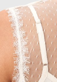 Durchsichtige Spitzenbluse mit gepunktetem Muster, versehen mit einem zarten Spitzenrand am Saum und einer weichen Textur. Helle Cremefarbe und verstellbare Träger.